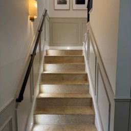 Renovest - Wykończenie Wnętrz - Eleganckie schody wyłożone beżowymi płytkami, oświetlone kinkietami i lampami sufitowymi, z drewnianą poręczą i panelami ściennymi w stylu klasycznym.