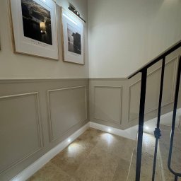 Renovest - Wykończenie Wnętrz - Eleganckie wykończenie ściany z panelami, oświetleniem punktowym przy podłodze i obrazami w ramkach. Widoczne fragmenty schodów z czarną balustradą.