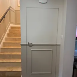 Ukryte drzwi w panelowej ścianie, dopasowane kolorystycznie do schod&oacute;w z trawertynu i parkietu w jodełkę. Minimalistyczny design, listwy przypodłogowe.