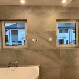 Renovest - Wykończenie Wnętrz - Elegancka łazienka z wanną, wykończona płytkami imitującymi marmur, z dwoma oknami z widokiem na budynek. Nowoczesna armatura, oświetlenie punktowe.