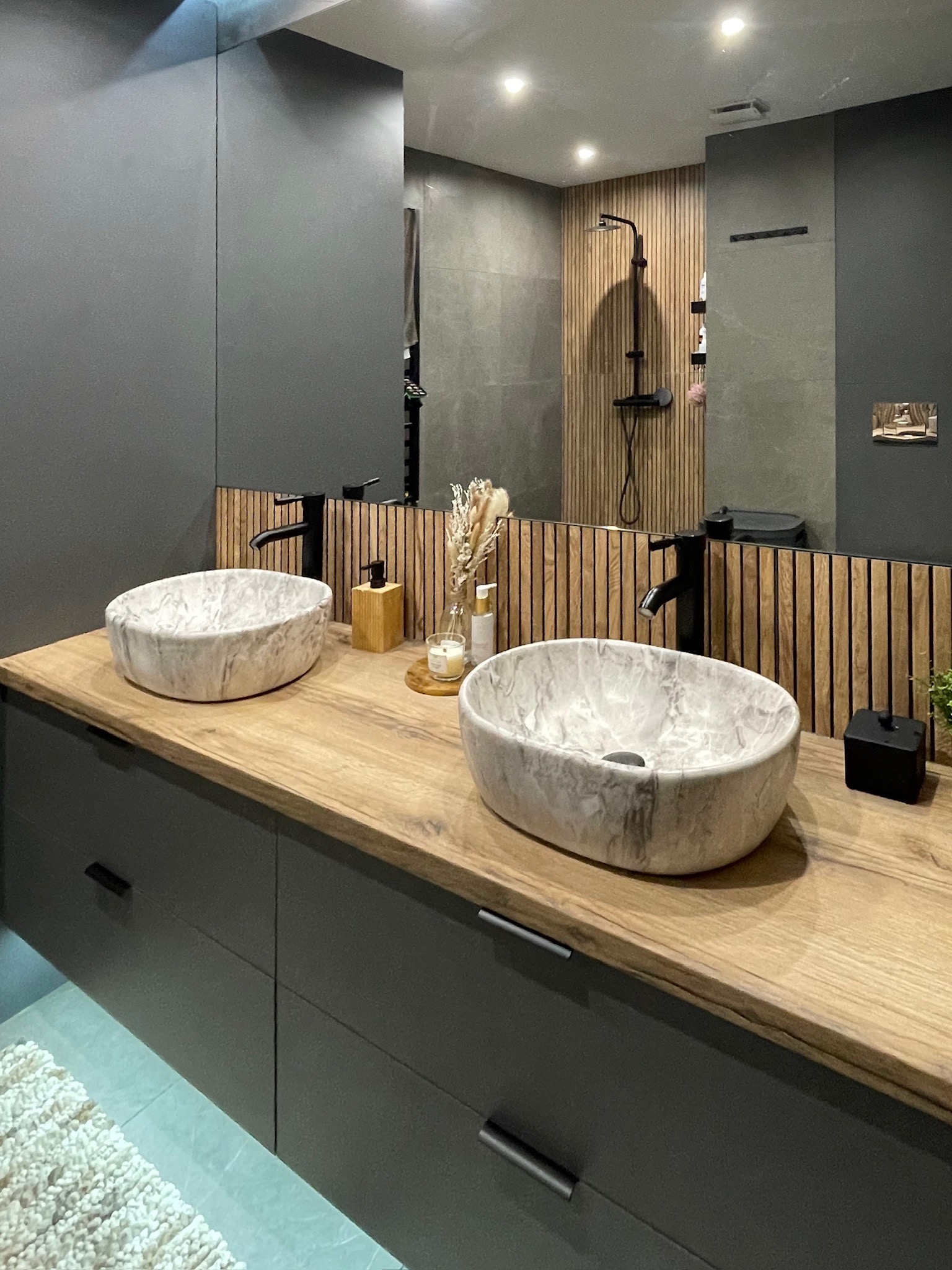 Nowoczesna łazienka z dwoma marmurkowymi umywalkami na drewnianym blacie i szarą zabudową. W tle lustro odbija prysznic z drewnianymi panelami.