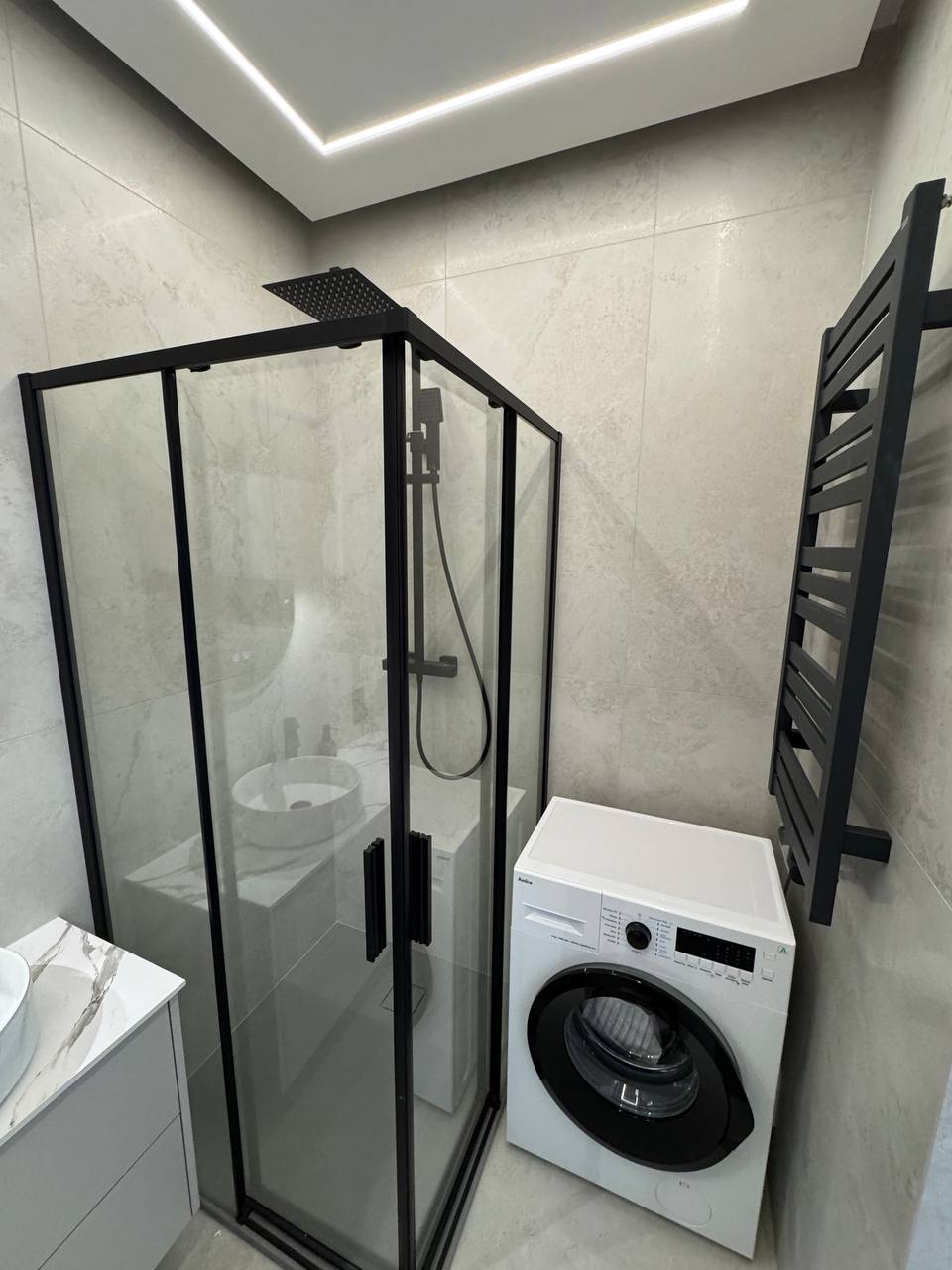 Nowoczesna łazienka w szarościach: kabina prysznicowa z czarnymi ramami, umywalka na blacie z marmuru, pralka i grzejnik drabinkowy. Minimalistyczny design.