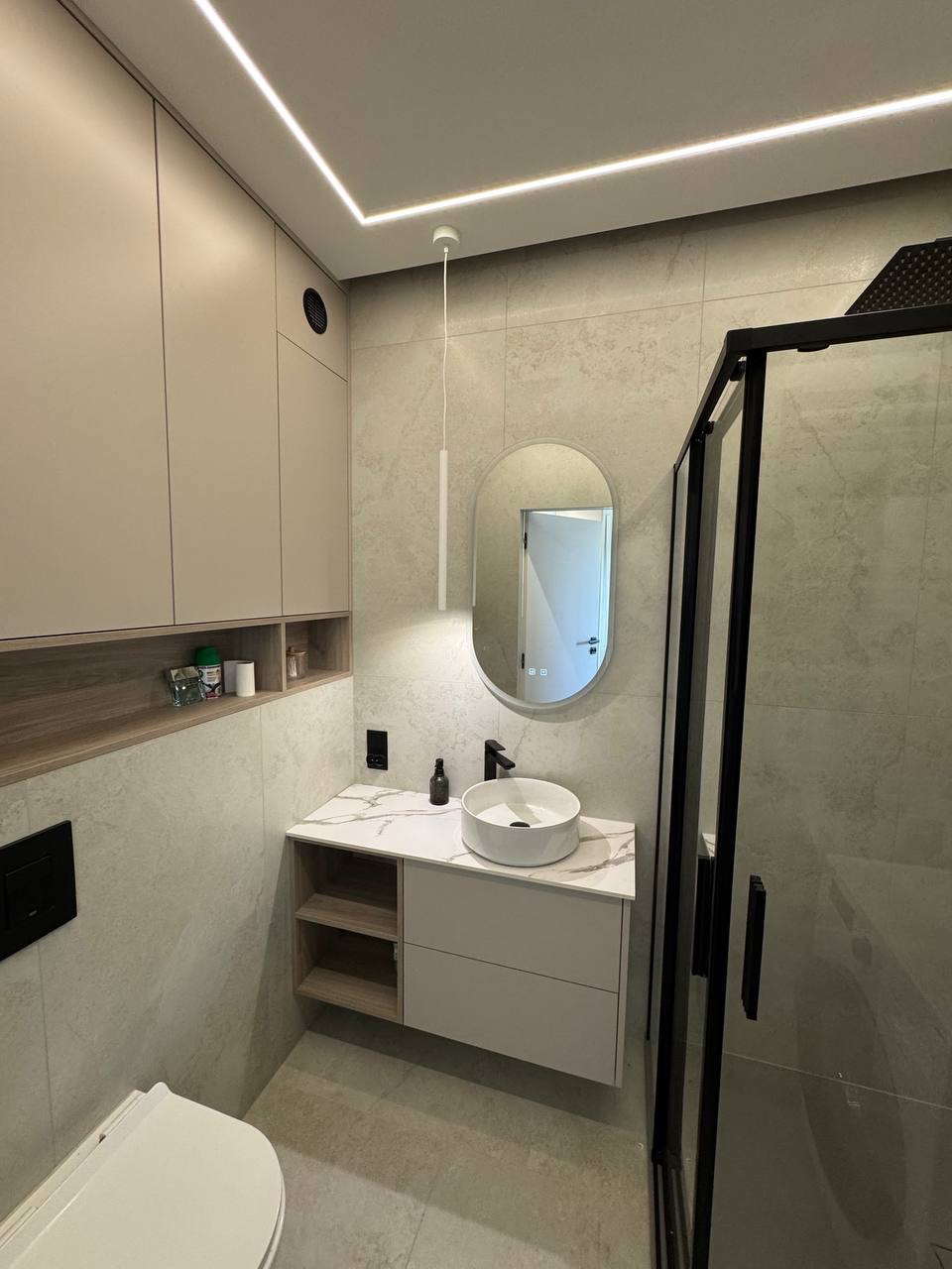 Nowoczesna łazienka z owalnym lustrem, umywalką nablatową i kabiną prysznicową z czarnymi profilami. Stonowana kolorystyka i oświetlenie LED.