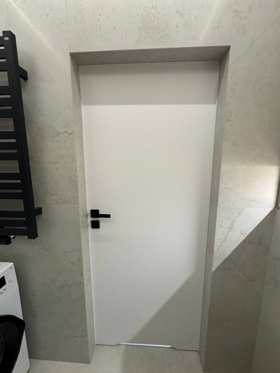 Białe, minimalistyczne drzwi z czarną klamką w łazience wykończonej płytkami imitującymi kamień. Widoczny fragment grzejnika i pralki.