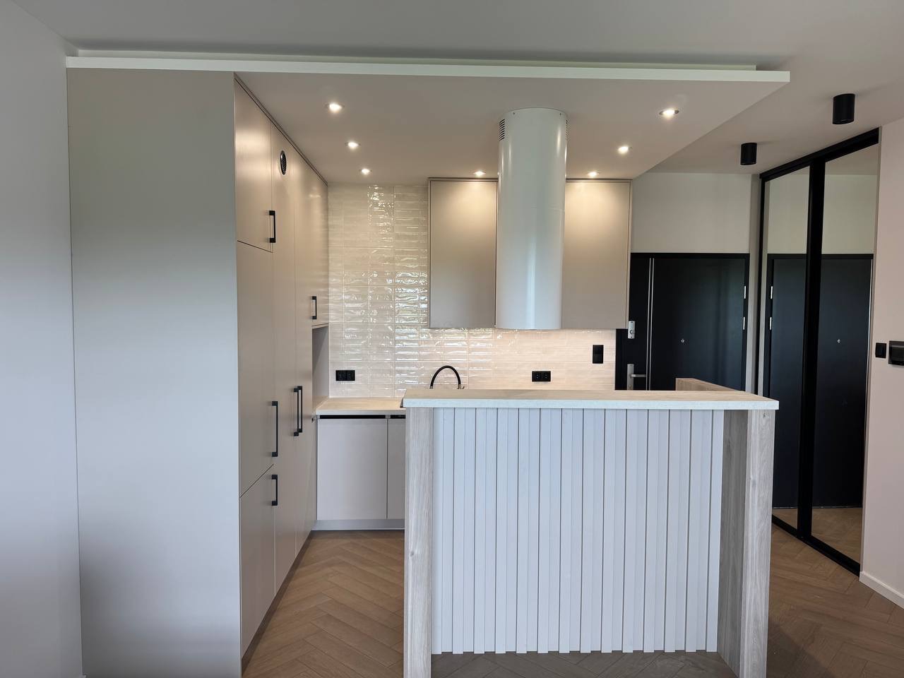 Jasna kuchnia z wyspą, płytki na ścianie, parkiet w jodełkę. Minimalistyczny design z cylindrycznym okapem i czarnymi akcentami.