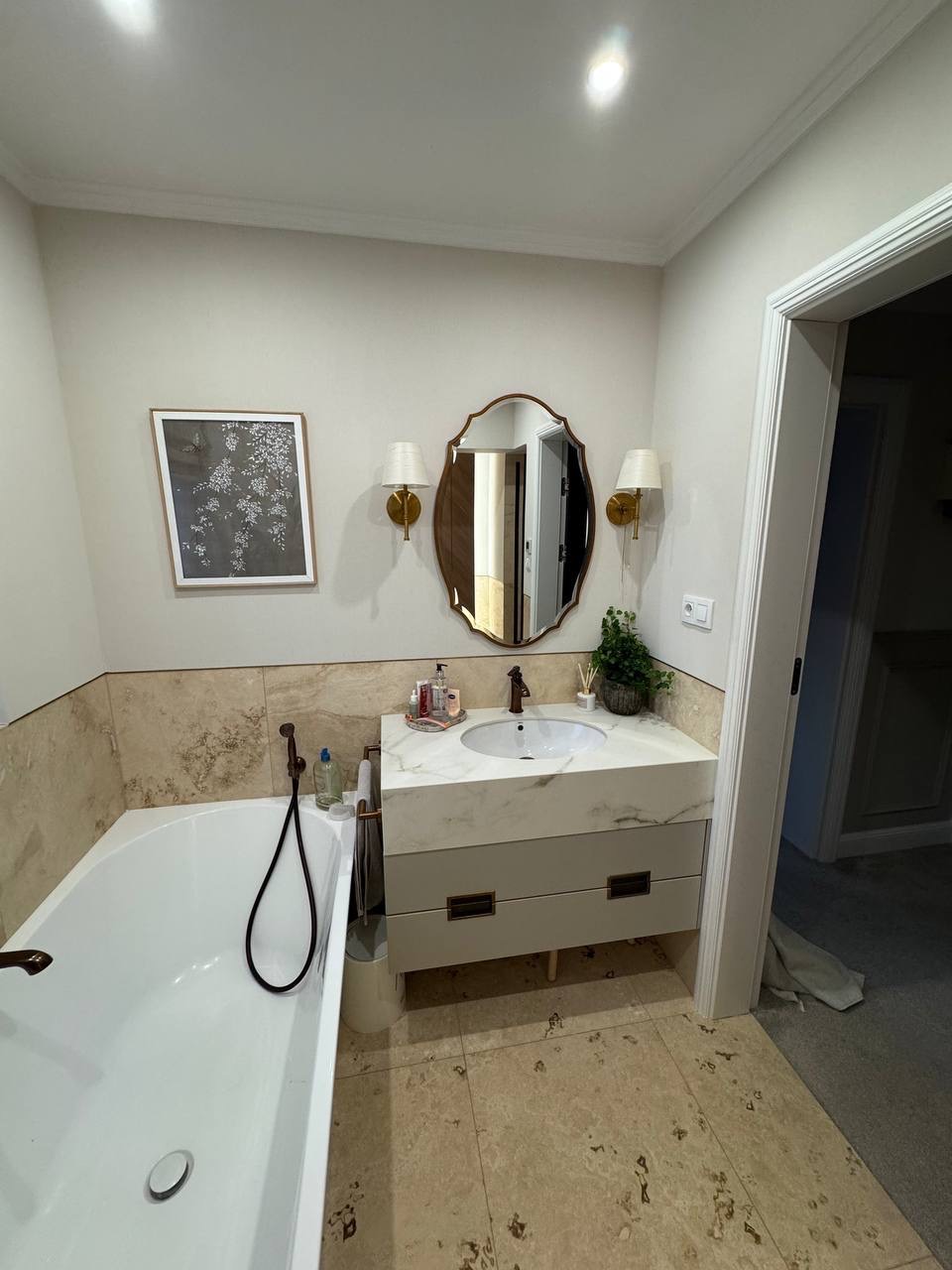 Elegancka łazienka z wanną, umywalką w marmurowym blacie i owalnym lustrem. Beżowe płytki i złote akcenty lamp tworzą spójny, luksusowy wystrój.