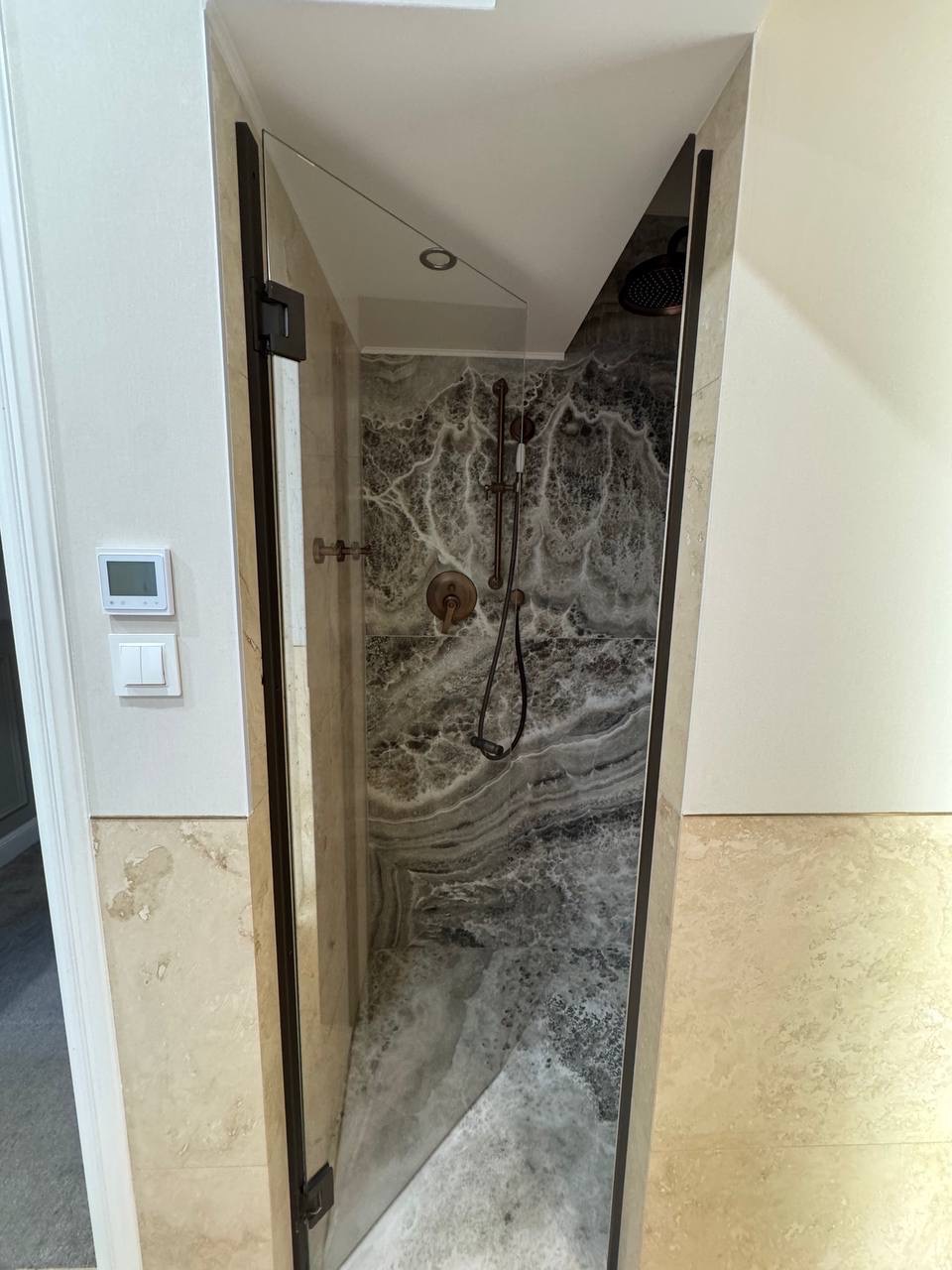 Narożna kabina prysznicowa z szarą, marmurkową okładziną, brązową armaturą i szklanymi drzwiami, wkomponowana w beżową łazienkę.