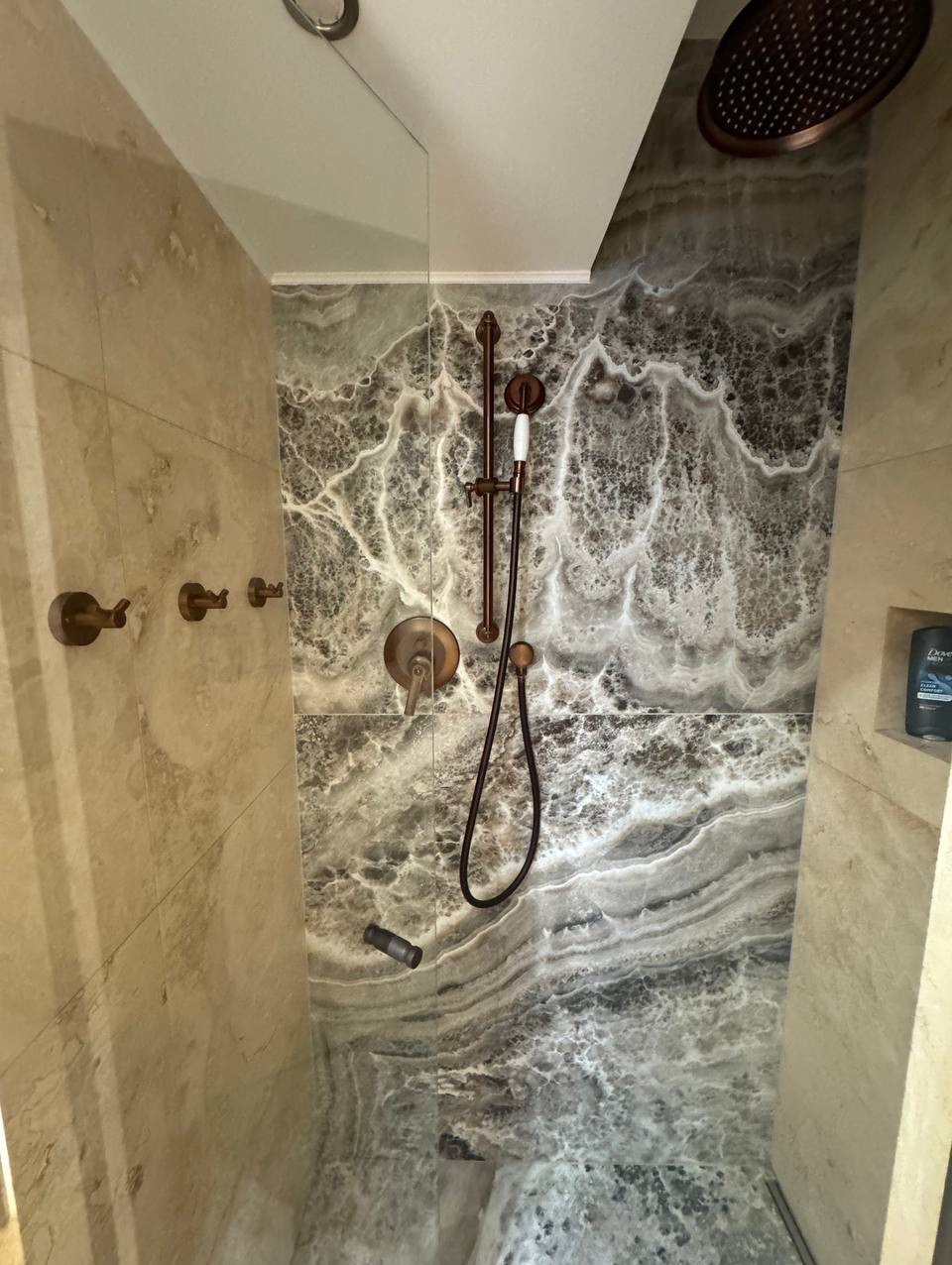 Elegancka kabina prysznicowa z marmurowymi płytkami w odcieniach szarości i beżu, z brązową armaturą i deszczownicą. Nowoczesny design.