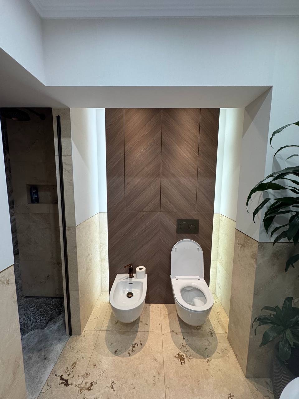 Nowoczesna łazienka z bidetem i toaletą podwieszaną, drewniane panele na ścianie, beżowe płytki podłogowe. Minimalistyczny design z rośliną doniczkową.