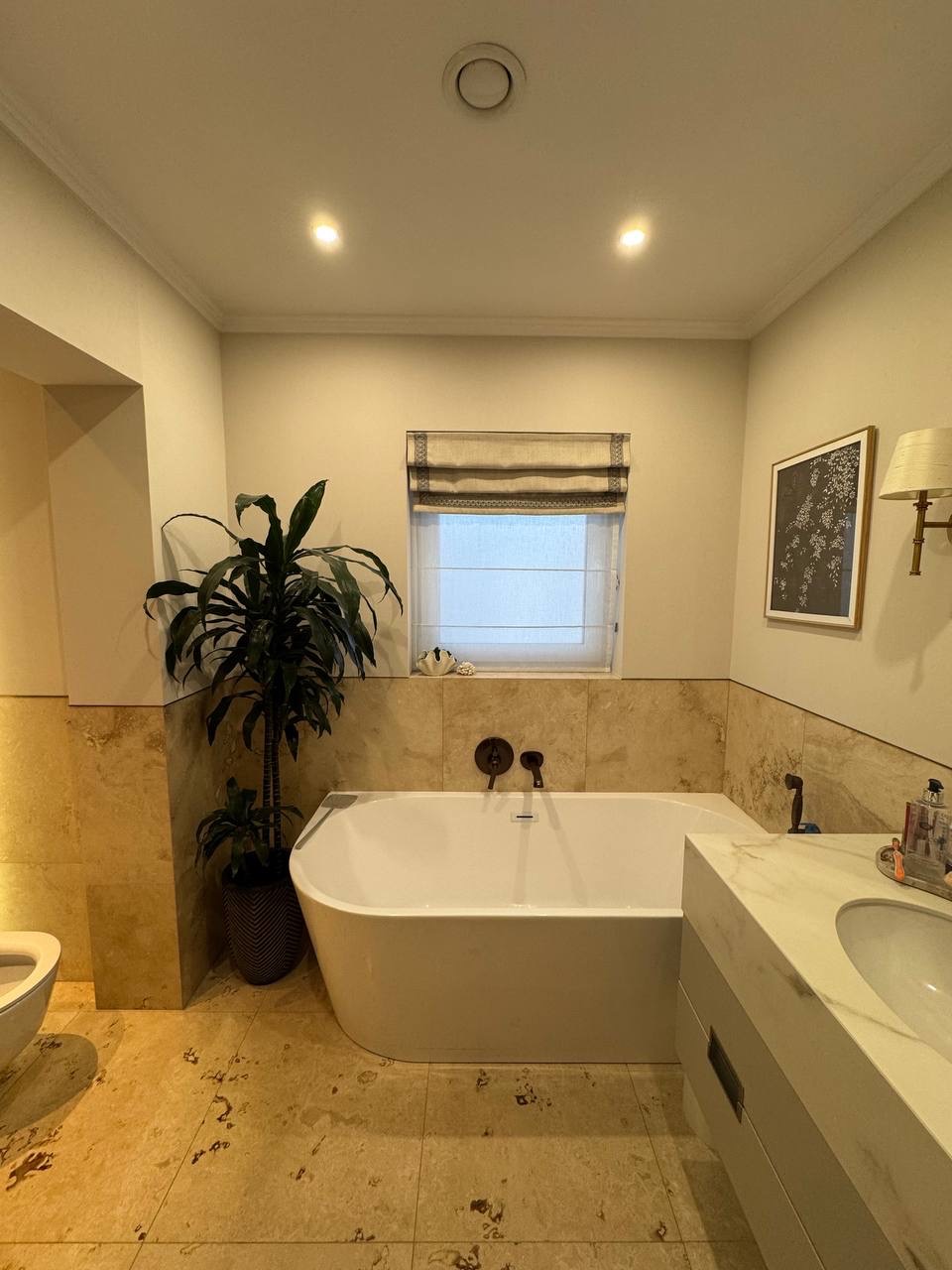 Jasna łazienka z wolnostojącą wanną, beżowymi płytkami, rośliną doniczkową i oknem z roletą rzymską. Armatura w kolorze starego złota.