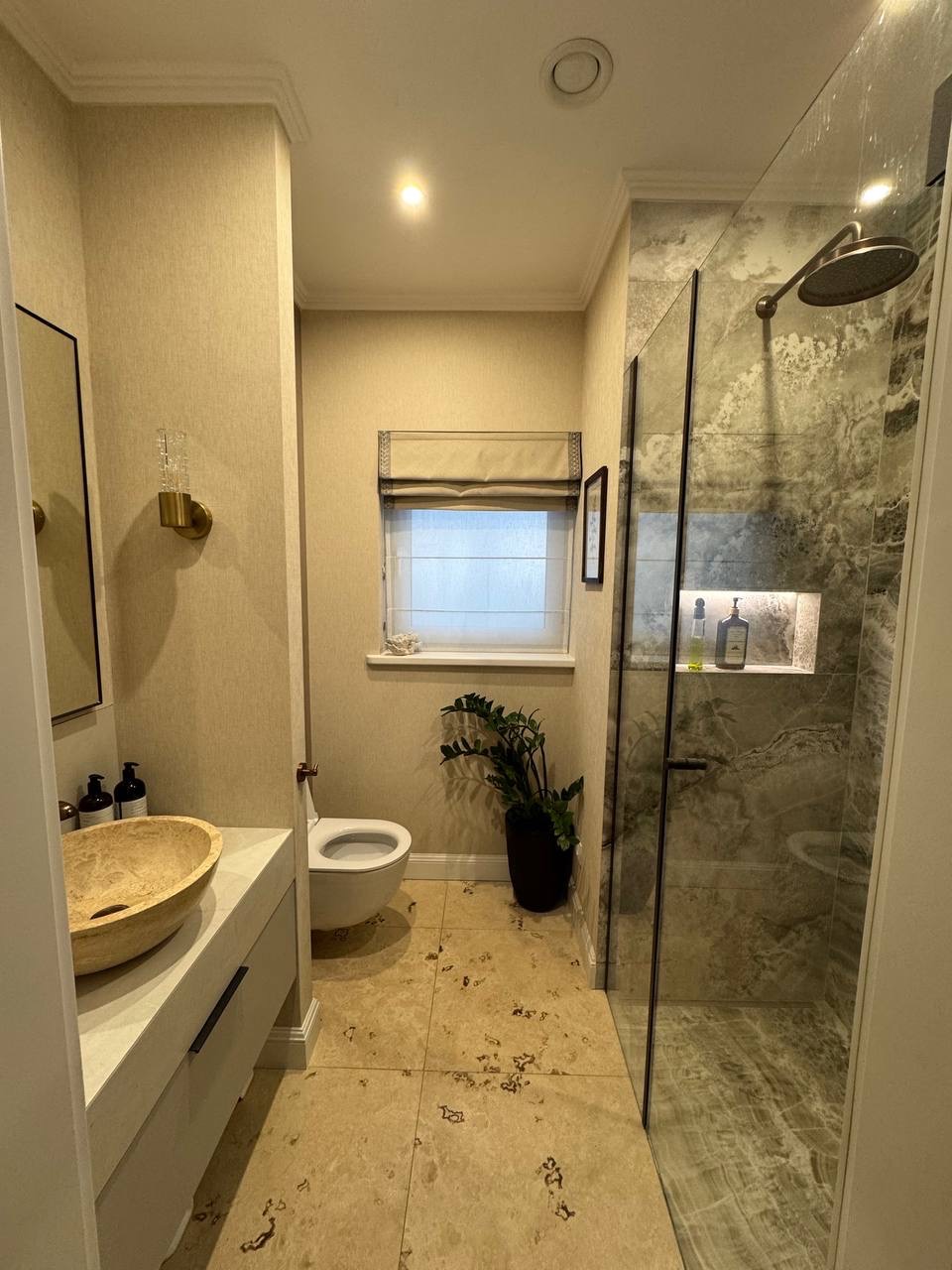 Jasna łazienka z beżowymi płytkami, umywalką z kamienia i kabiną prysznicową z marmurowymi ścianami. Uwagę zwraca roślina w czarnej donicy pod oknem.