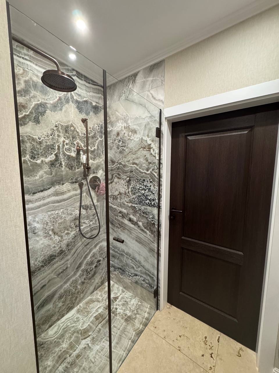 Kabina prysznicowa z marmurowymi płytami i deszczownicą, widoczna obok ciemna drewniana stolarka drzwiowa, dopełnia aranżację łazienki.