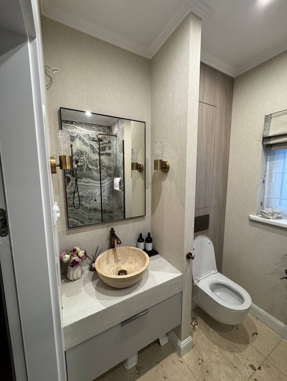 Elegancka łazienka z beżową umywalką w kształcie misy, lustrem z widokiem na prysznic i toaletą. Neutralna kolorystyka z akcentami złota.