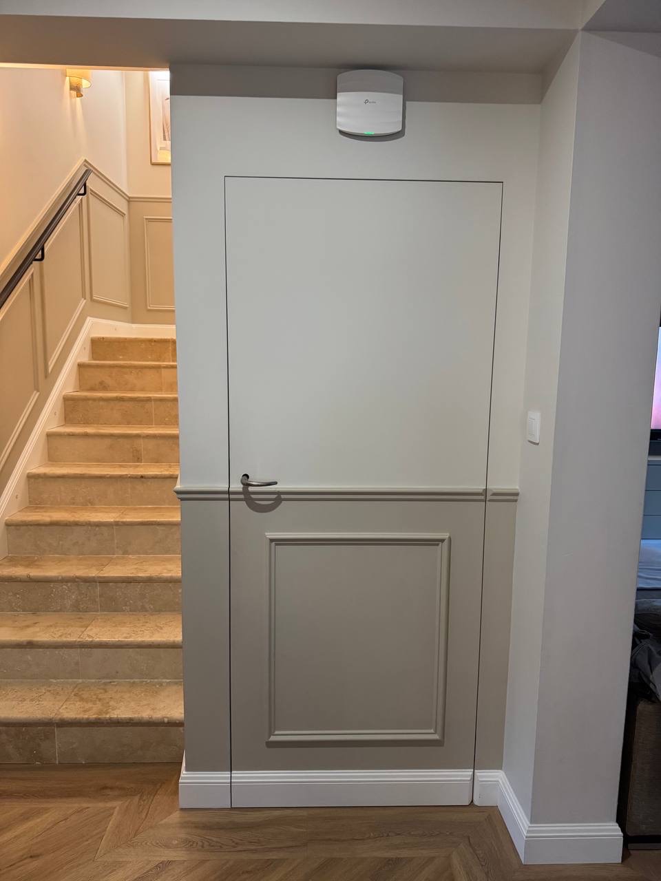 Ukryte drzwi w panelowej ścianie, dopasowane kolorystycznie do schodów z trawertynu i parkietu w jodełkę. Minimalistyczny design, listwy przypodłogowe.