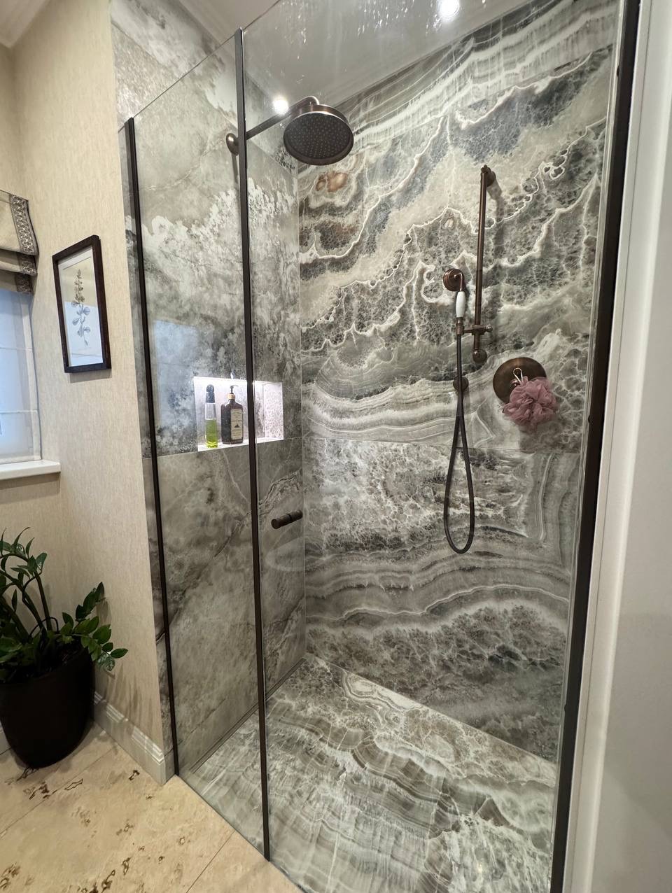 Kabina prysznicowa z marmurowymi ścianami i szklanymi drzwiami, widok na baterię w kolorze brązu i wnękę z kosmetykami. Eleganckie wykończenie łazienki.