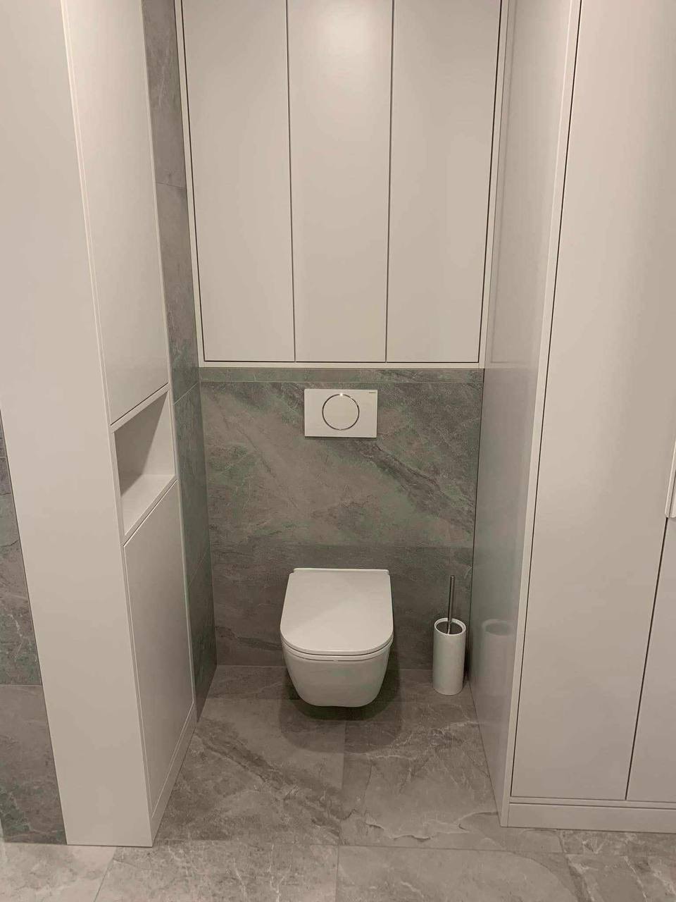 Nowoczesna, minimalistyczna łazienka z podwieszaną toaletą, szarymi płytkami imitującymi marmur i białymi szafkami w zabudowie. Widoczny szczotka do WC i przycisk spłuczki.