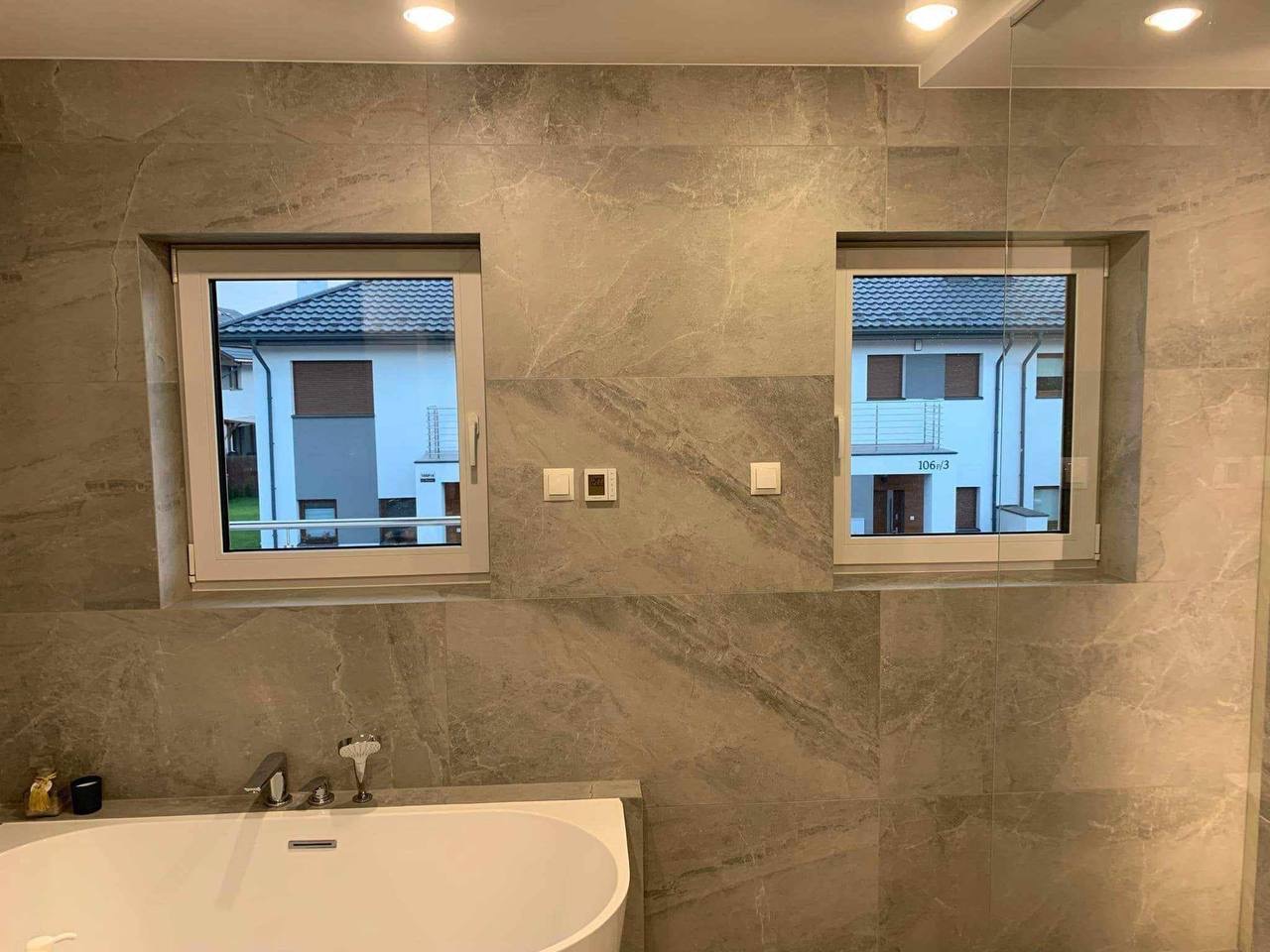 Elegancka łazienka z wanną, wykończona płytkami imitującymi marmur, z dwoma oknami z widokiem na budynek. Nowoczesna armatura, oświetlenie punktowe.