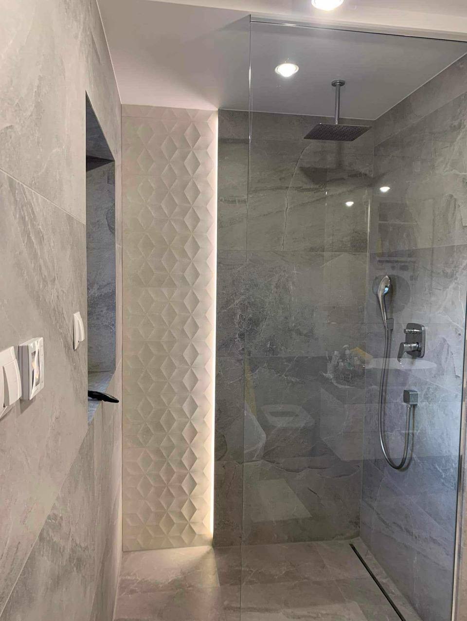 Nowoczesna łazienka z kabiną prysznicową typu walk-in, szare płytki imitujące marmur i dekoracyjna ściana z podświetlonymi, trójwymiarowymi panelami.