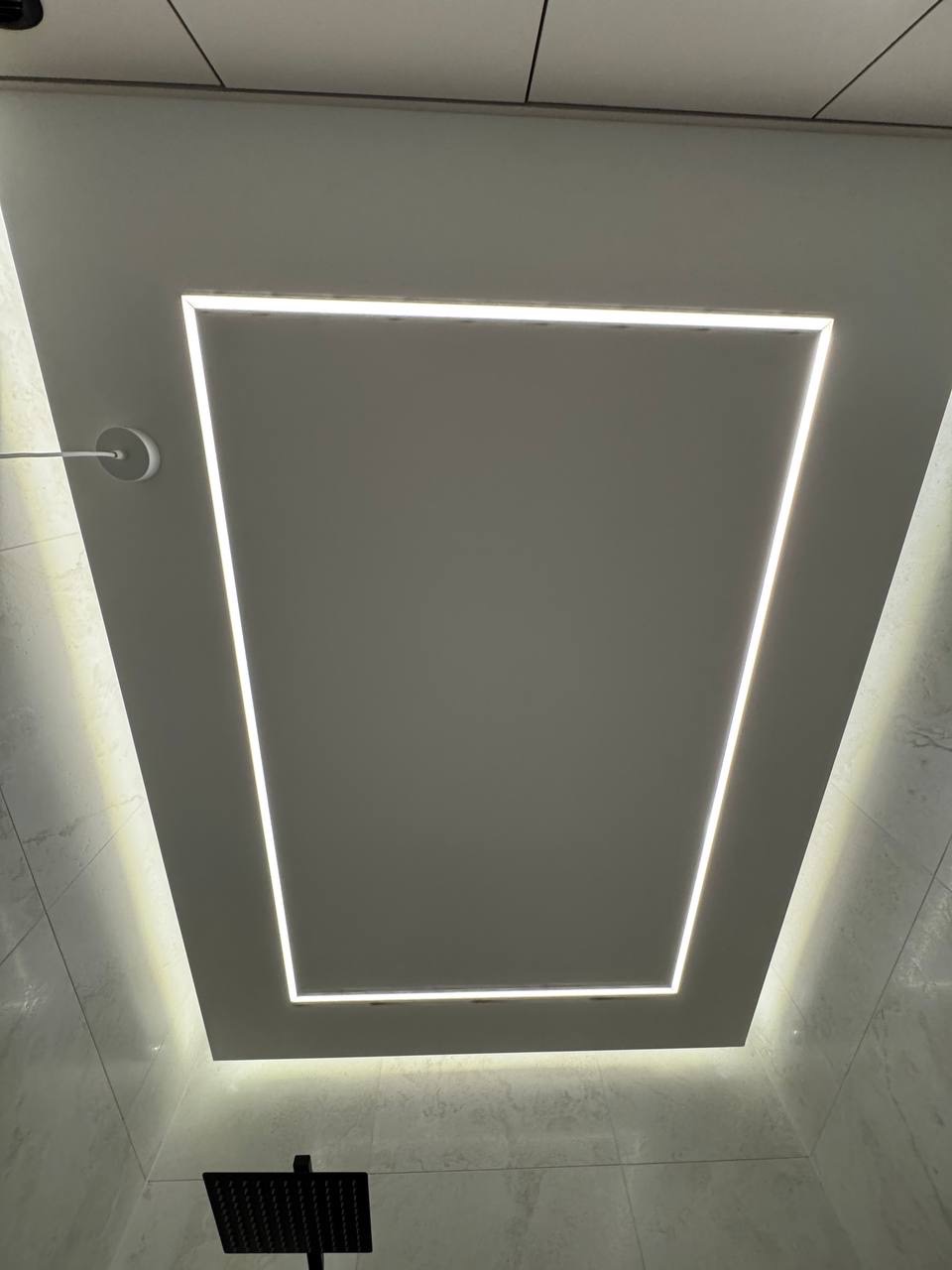 Nowoczesna łazienka z podświetlanym sufitem w kształcie kwadratu. Białe płytki na ścianach. Czarna deszczownica. Minimalistyczny design z efektem subtelnego oświetlenia.