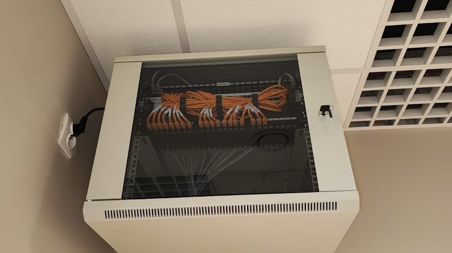 Zamknięta szafa rack z pomarańczowymi kablami sieciowymi i patch panelem, zamontowana na ścianie w pomieszczeniu biurowym z kratownicowym sufitem podwieszanym.