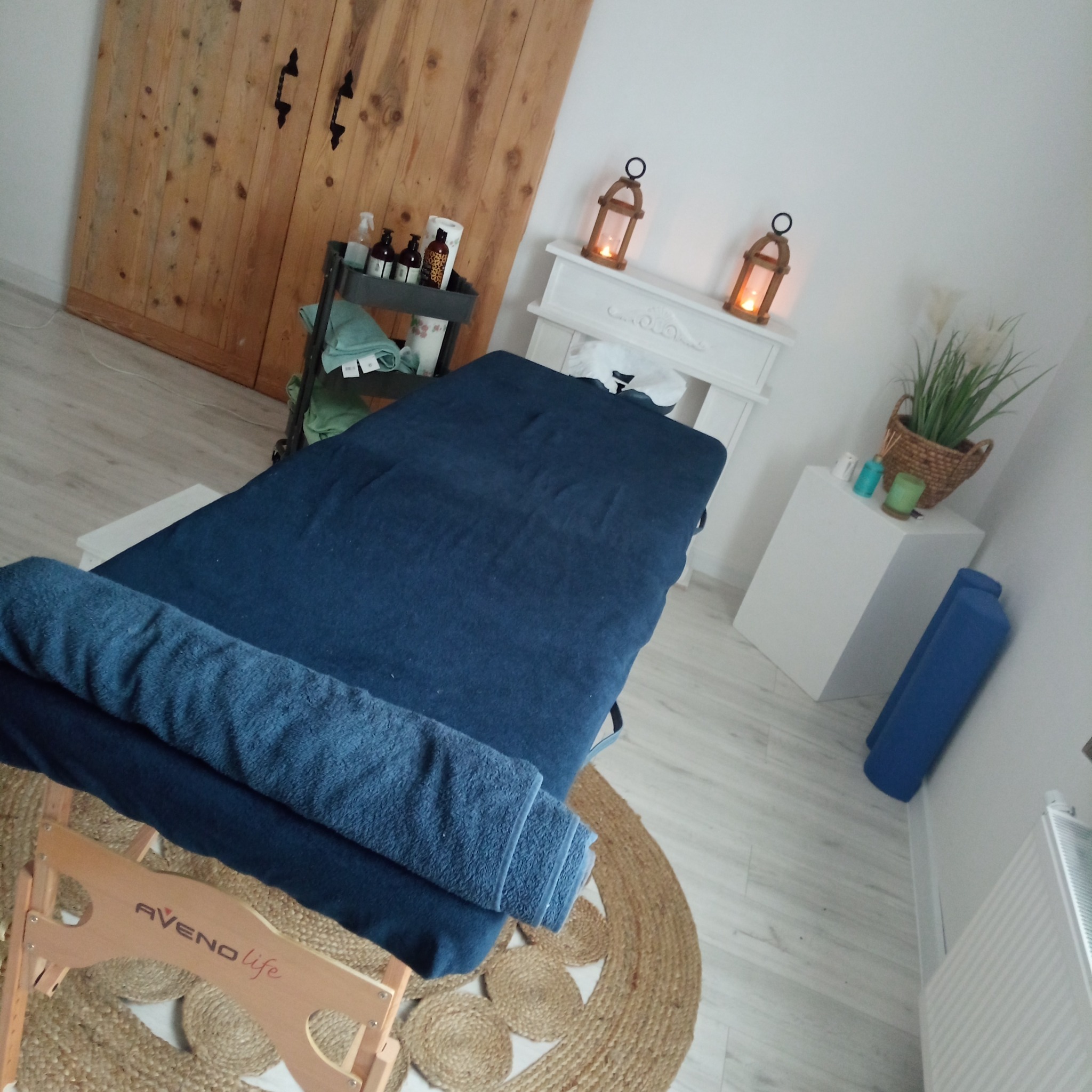 Stół do zabiegów z ciemnoniebieskim pokrowcem i ręcznikami w przytulnym gabinecie, oświetlonym lampionami. Na podłodze leży okrągły, jutowy dywan.