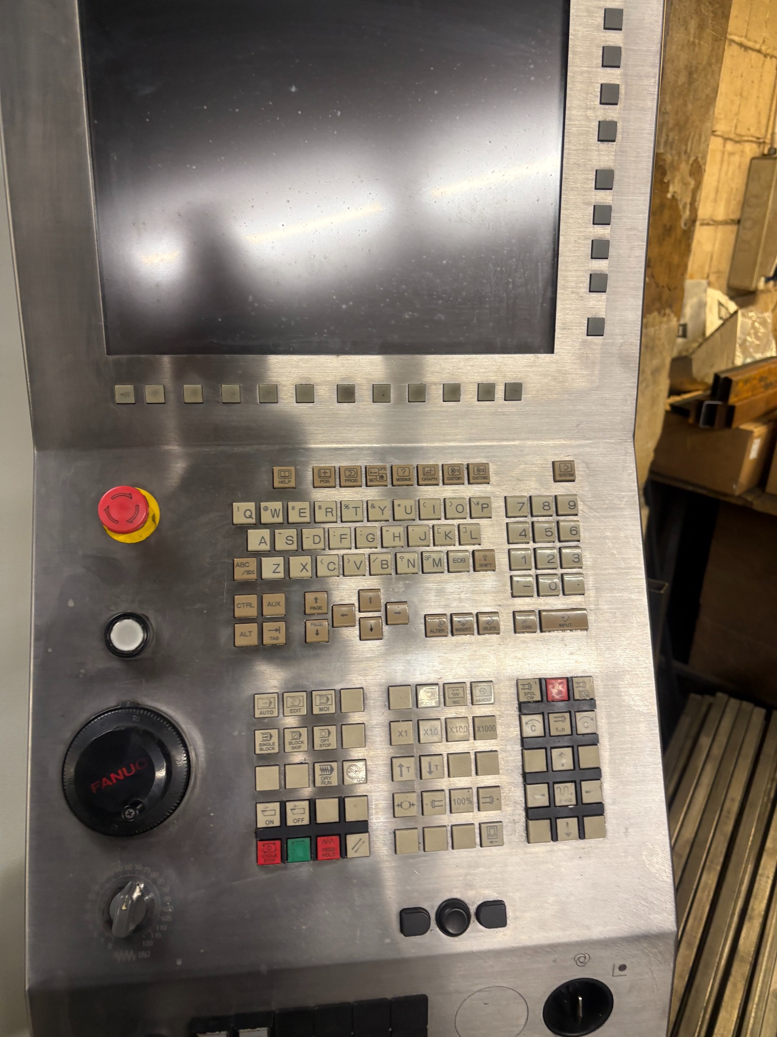 Panel sterowania numerycznej maszyny CNC marki Fanuc z widocznym ekranem, klawiaturą alfanumeryczną i przyciskami funkcyjnymi. Metalowa obudowa, przycisk bezpieczeństwa.