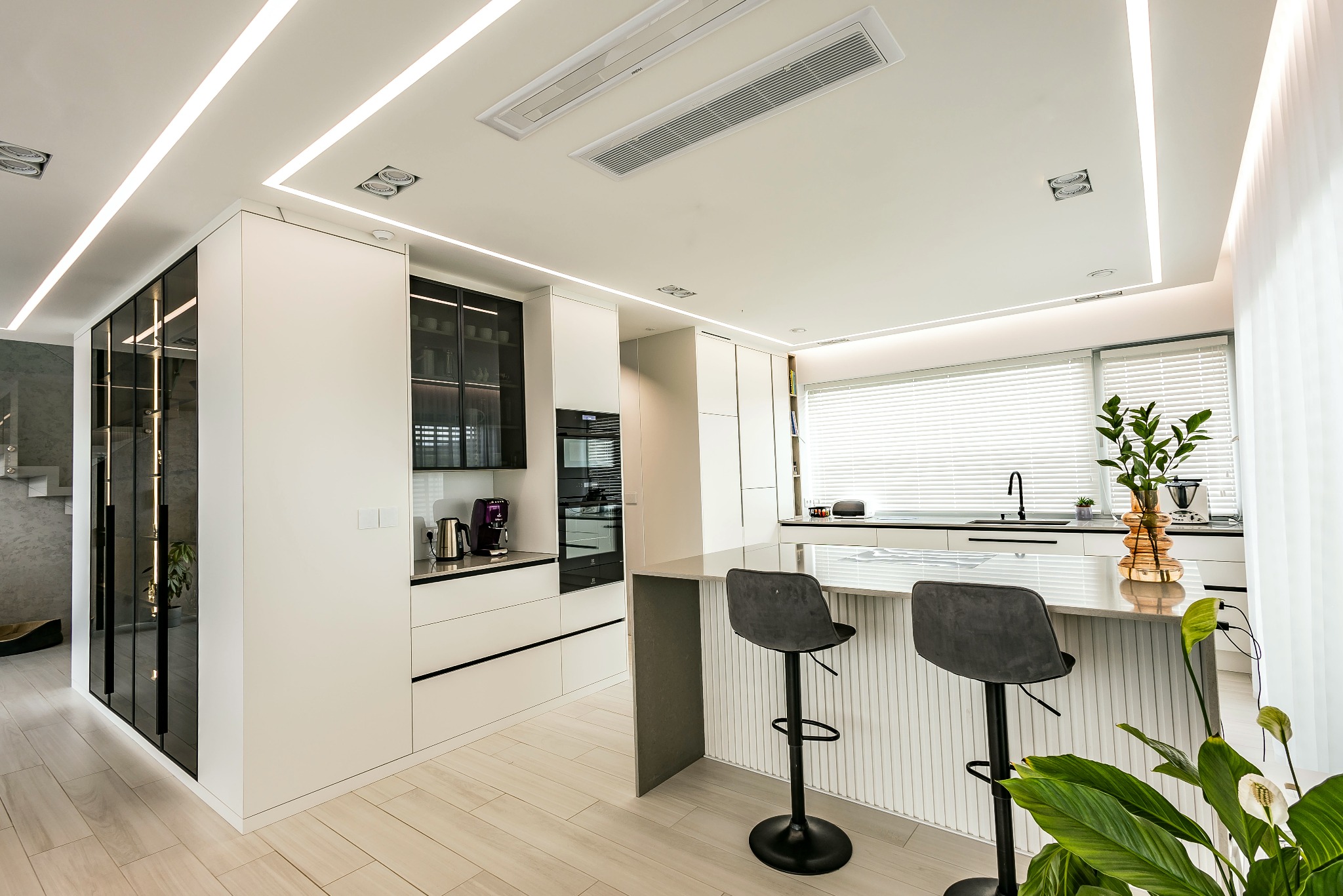 Nowoczesna, jasna kuchnia z wyspą i dwoma hokerami. Białe szafki z czarnymi akcentami, drewniana podłoga, oświetlenie LED i rośliny doniczkowe dodają elegancji.