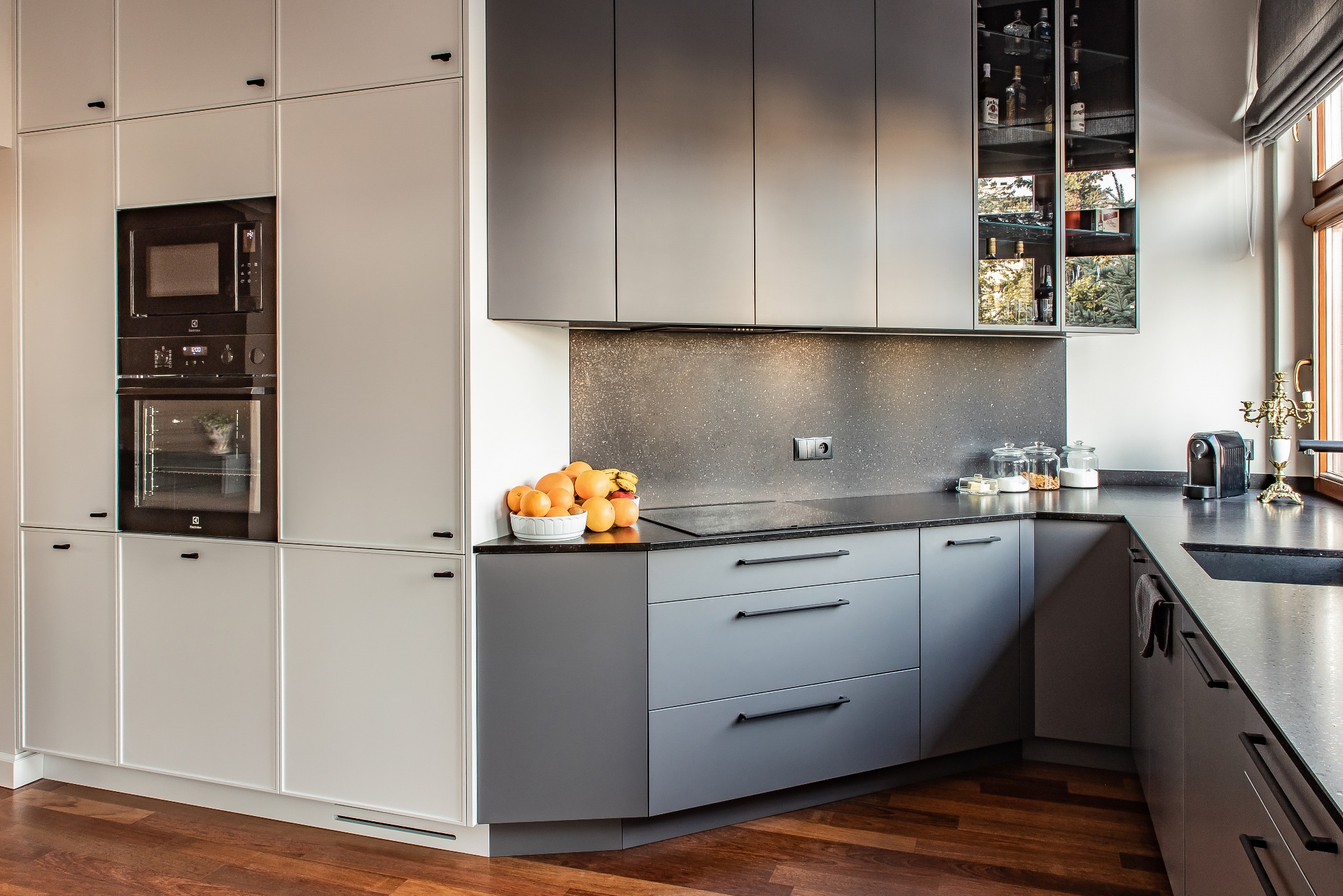 Nowoczesna kuchnia w szaro-białej tonacji z drewnianą podłogą. Zabudowa kuchenna z piekarnikiem Electrolux i szafką z przeszklonymi drzwiami. Owoce na blacie.