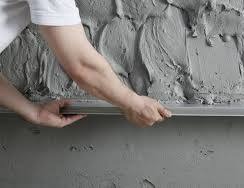 Ręka wygładza szpachlą ścianę z nierównomiernie nałożoną, szarą masą, widoczna część ramienia i narzędzie. Detal wykończenia wnętrza.