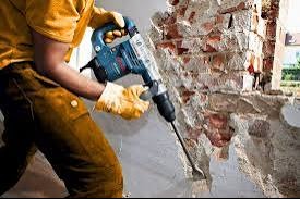 Kucie ściany z cegły wewnątrz budynku za pomocą młota udarowego, pracownik w rękawicach ochronnych. Widoczne fragmenty instalacji elektrycznej.