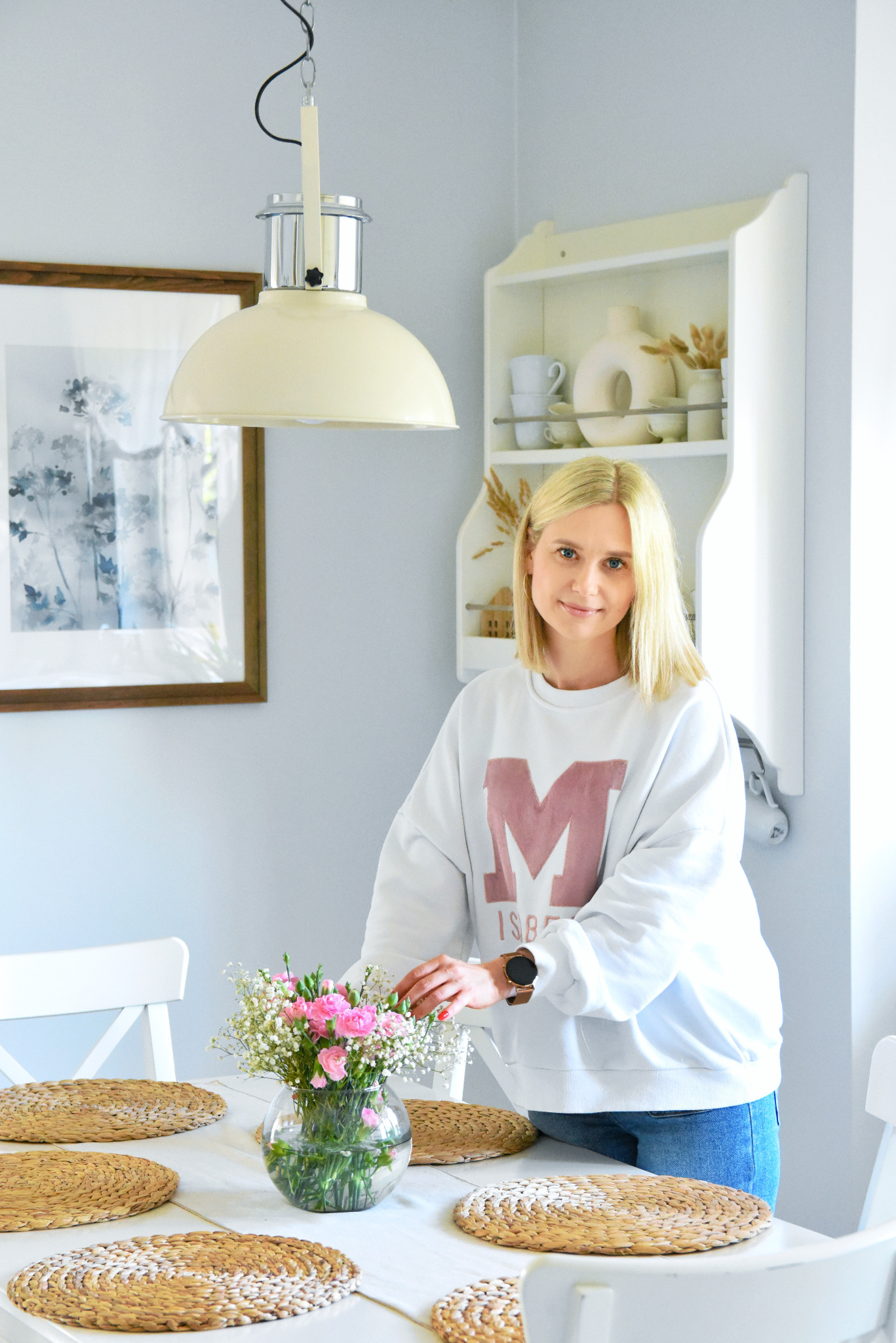 Jasna jadalnia ze stołem i blondynką układającą kwiaty w wazonie; nad stołem lampa w stylu retro, w tle półka z dekoracjami i obraz w drewnianej ramie.