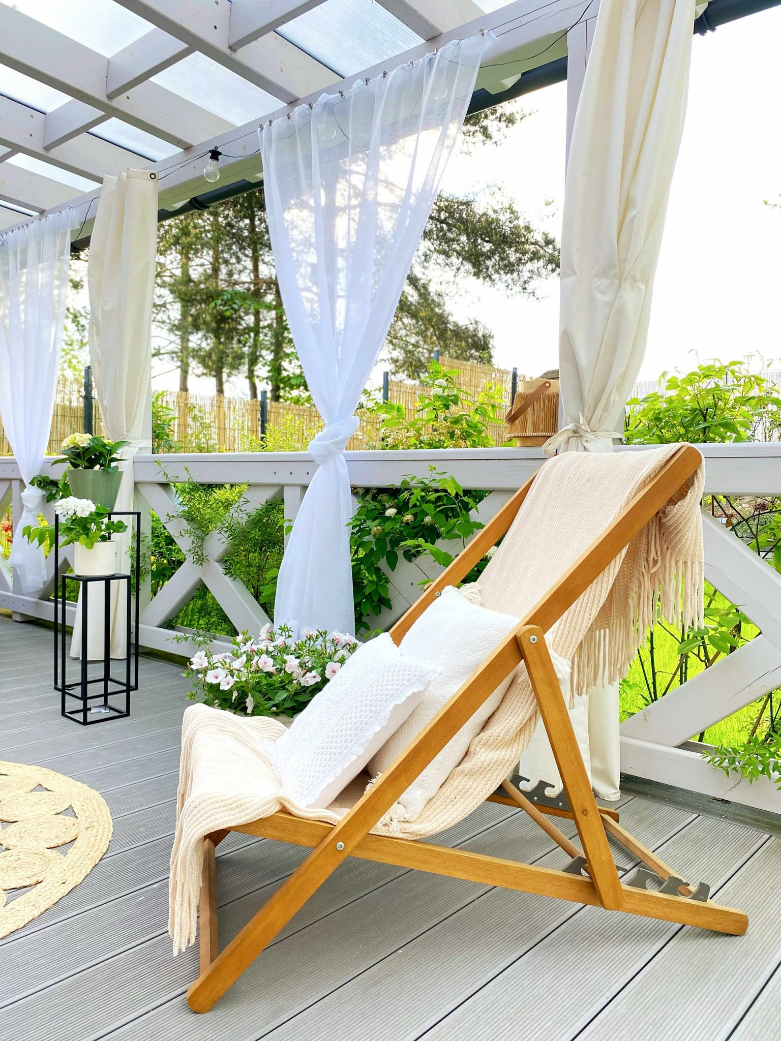 Jasny taras z drewnianym leżakiem, białymi poduszkami i pledem. W tle zwiewne zasłony, rośliny i dekoracje tworzą przytulną strefę relaksu.