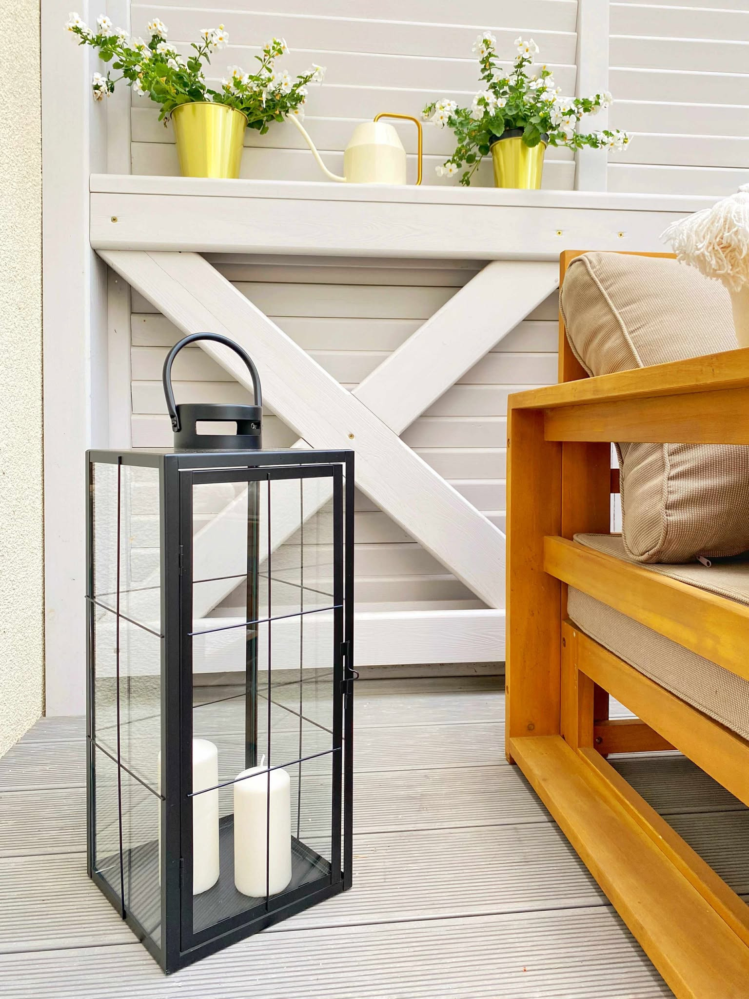 Aranżacja balkonu z drewnianym krzesłem z poduszkami i czarnym lampionem ze świecami. Biała, drewniana ściana z dekoracjami kwiatowymi w złotych doniczkach.