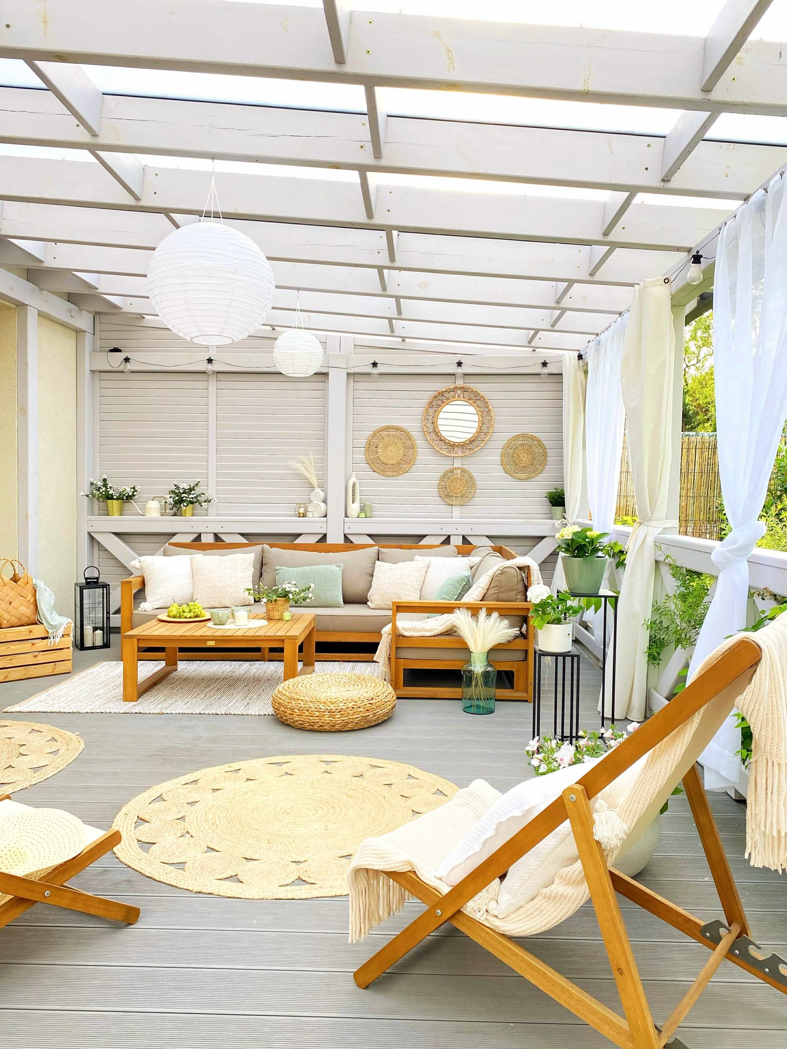 Zadaszony taras z drewnianymi meblami, okrągłymi dywanami z juty i białymi lampionami. Dekoracje w stylu boho, wiszące zasłony i rośliny doniczkowe.
