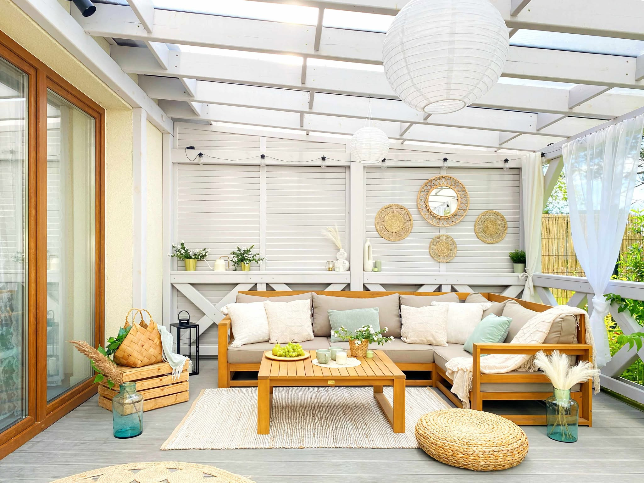 Stylowa aranżacja zadaszonego tarasu z drewnianym narożnikiem, stolikiem kawowym i dekoracjami w stylu boho. Jasne kolory, naturalne materiały, lampiony i tekstylia.