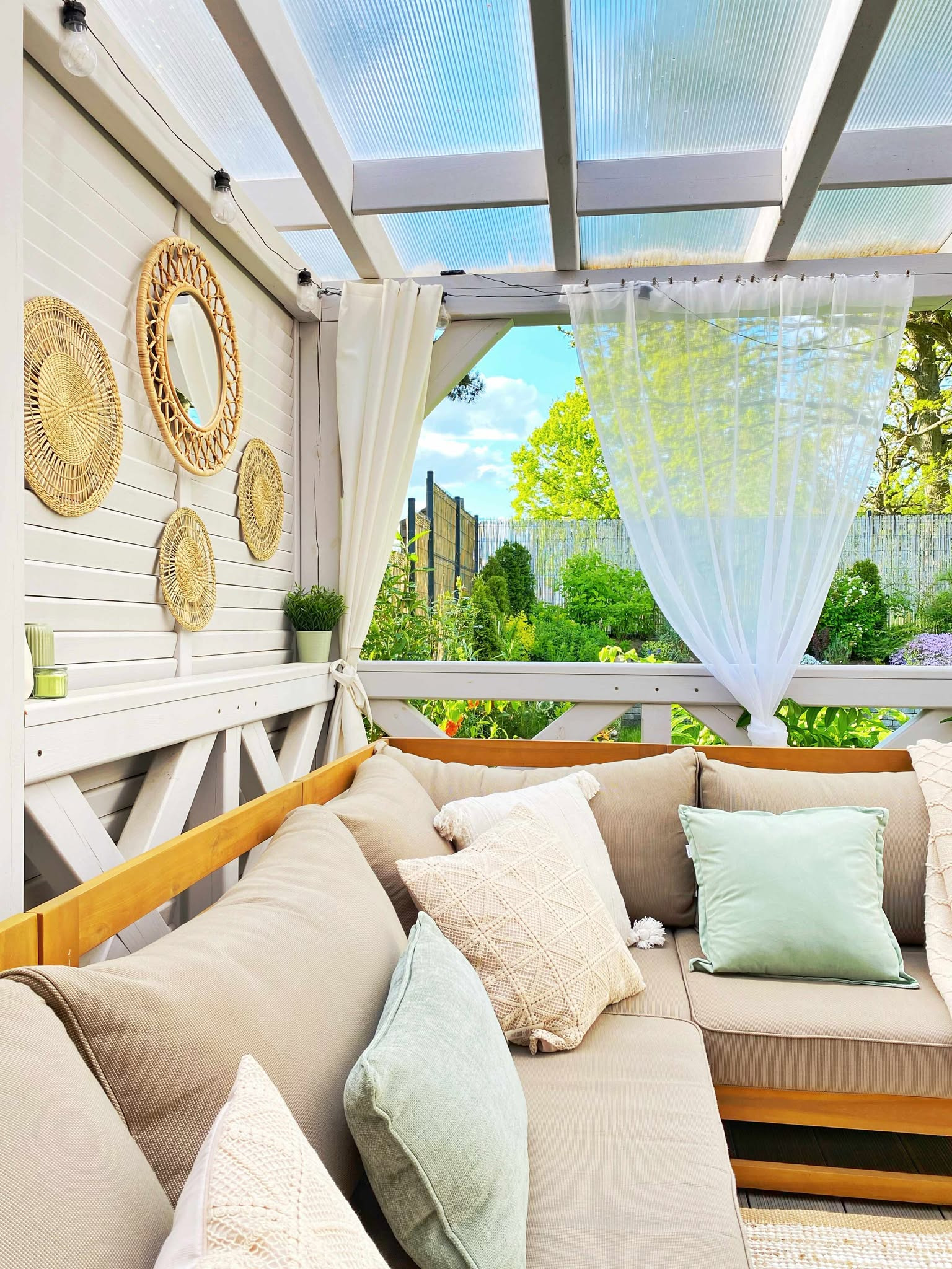 Jasny, zadaszony taras z beżową sofą, dekoracjami z wikliny i zwiewnymi zasłonami. Widok na zielony ogród w słoneczny dzień.