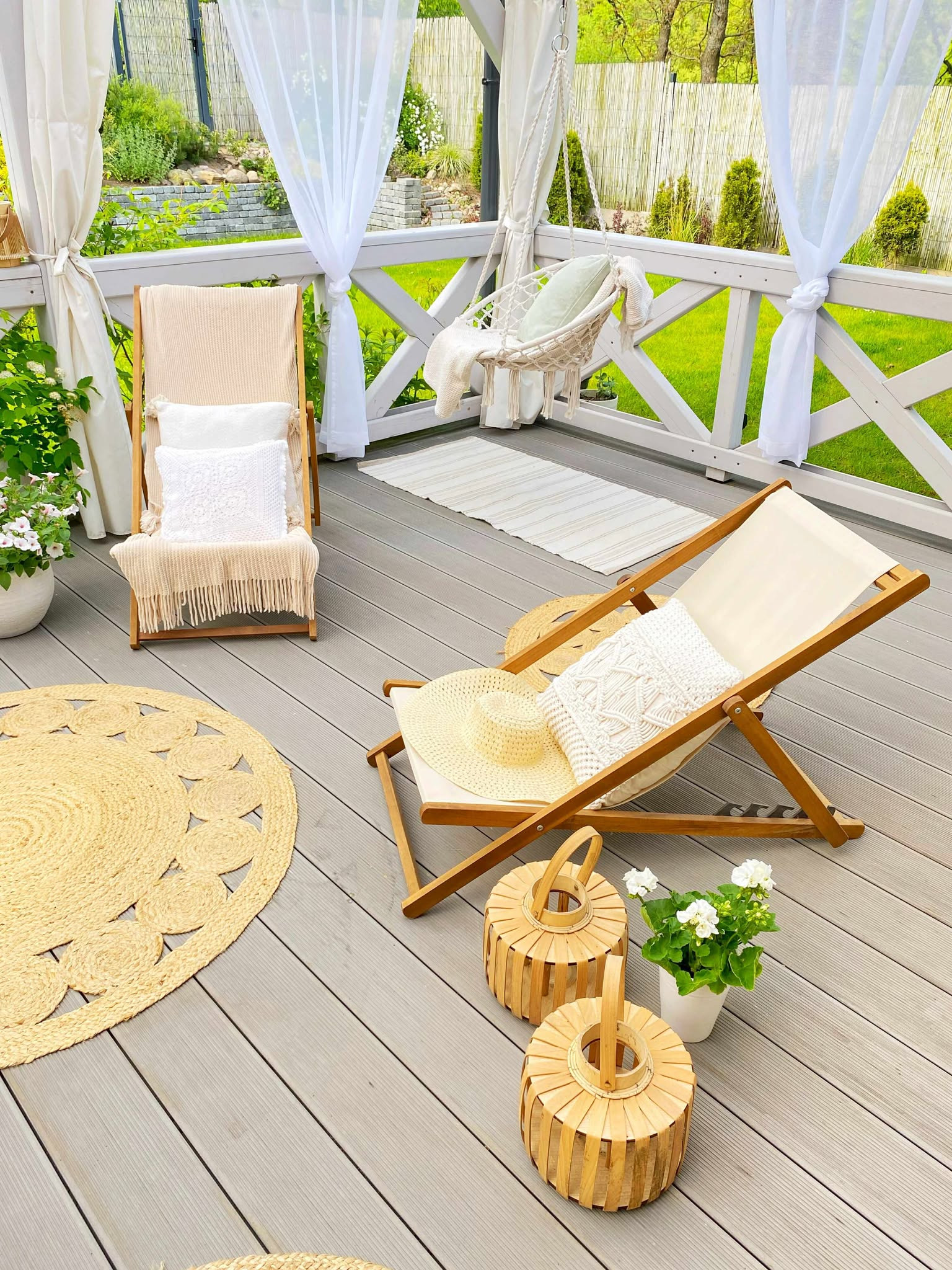 Balkon z drewnianą podłogą, leżakami, dywanem z juty i wiszącym fotelem, udekorowany białymi zasłonami i kwiatami w donicach. Letnia aranżacja.