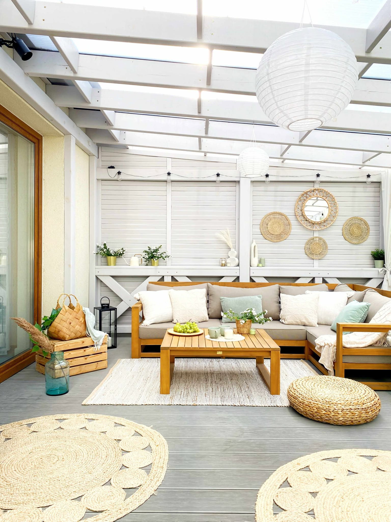Jasny, zadaszony taras z drewnianym narożnikiem, stolikiem i dywanami z juty. Dekoracje ścienne z wikliny i lampiony. Styl boho, naturalne materiały, relaksująca przestrzeń.