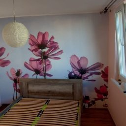 Arkadiusz Arko-Rem - Sypialnia po remoncie: ściana z fototapetą w kwiaty, drewniane łóżko z listwami, okno z widokiem na zieleń. Na parapecie akcesoria malarskie.