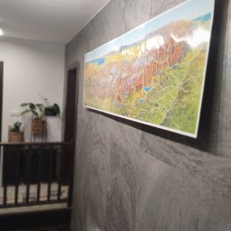 Arkadiusz Arko-Rem - Długi, panoramiczny obraz z mapą krajobrazową wisi na szarej, teksturowanej ścianie. W tle widoczne schody i rośliny doniczkowe, tworząc nowoczesne wnętrze.