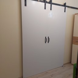 Arkadiusz Arko-Rem - Białe, przesuwne drzwi na czarnej, metalowej szynie z uchwytami, zamontowane w beżowym wnętrzu. Widoczna podłoga i fragment szafy.