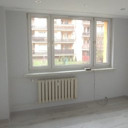 Arkadiusz Arko-Rem - Pomalowane na biało puste pomieszczenie z grzejnikiem pod oknem. Świeżo wykończone ściany i jasna podłoga. Widok na budynek zza okna.