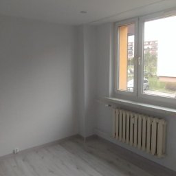 Arkadiusz Arko-Rem - Jasne, świeżo wyremontowane wnętrze z panelami podłogowymi, białymi ścianami i grzejnikiem pod oknem. Widok na zewnątrz przez okno.