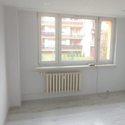 Arkadiusz Arko-Rem - Jasne, odnowione wnętrze pokoju z białymi ścianami, panelami podłogowymi i grzejnikiem pod oknem. Widok na budynek zza okna.