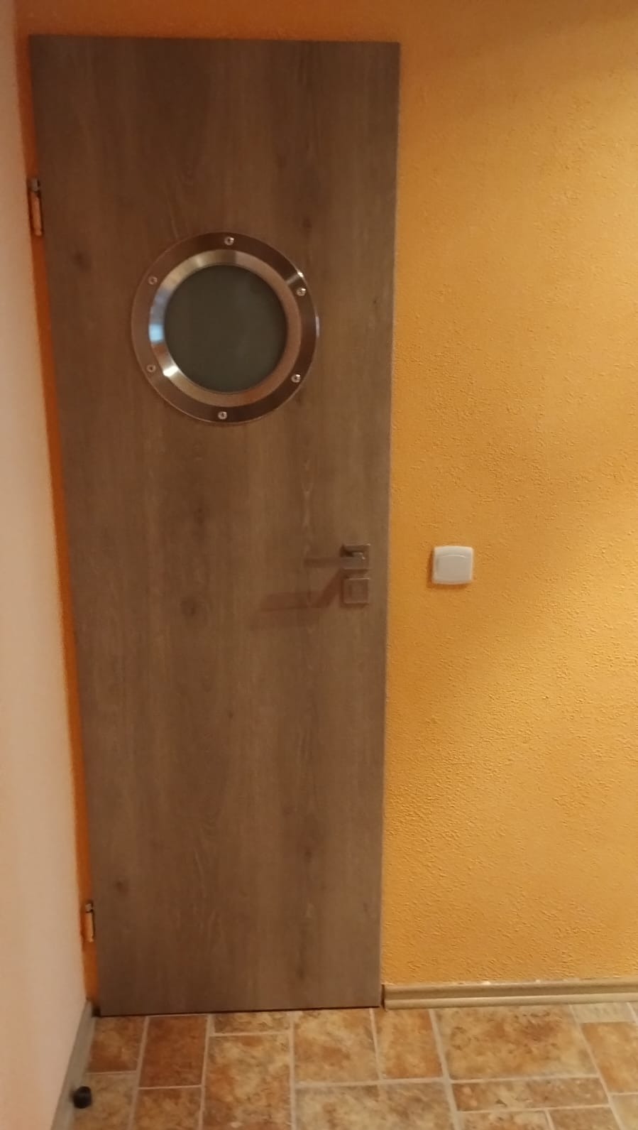 Zamknięte drzwi w stylu marynistycznym z okrągłym okienkiem w metalowej ramie, zamontowane w pomarańczowej ścianie, widoczna podłoga z płytek.