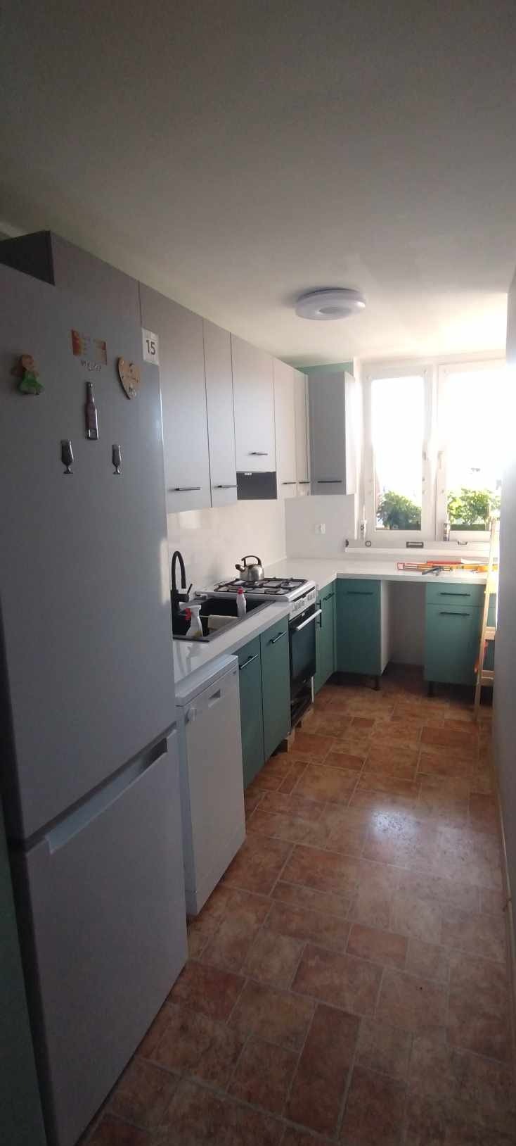 Jasna kuchnia po remoncie w Rybniku: białe szafki górne, turkusowe dolne, brązowa podłoga. Widok na okno i sprzęty AGD. Nowoczesny design i funkcjonalność.