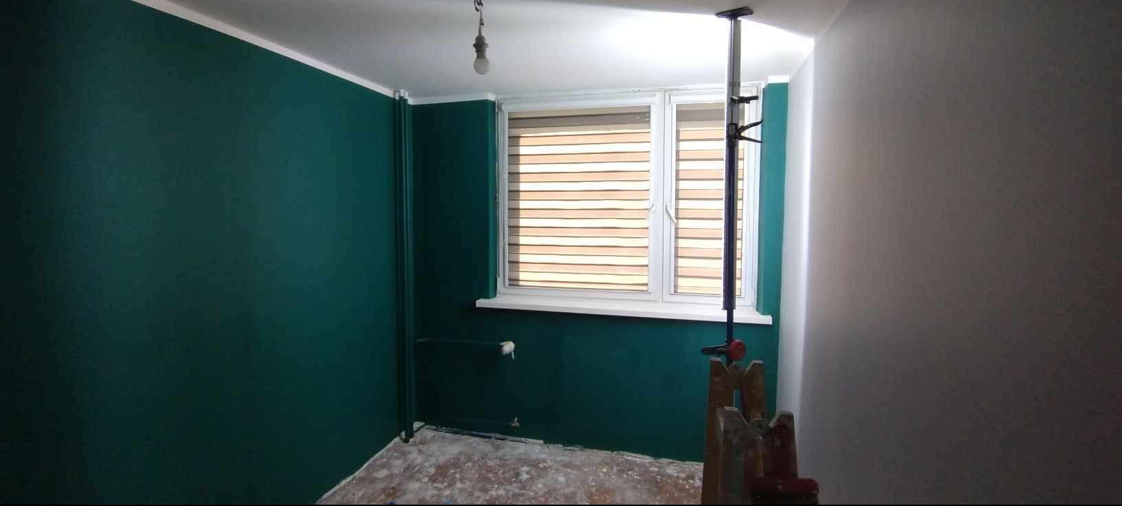 Pomieszczenie po malowaniu ścian na kolor butelkowej zieleni, widoczny grzejnik i okno z żaluzjami. Podłoga zabezpieczona folią malarską.
