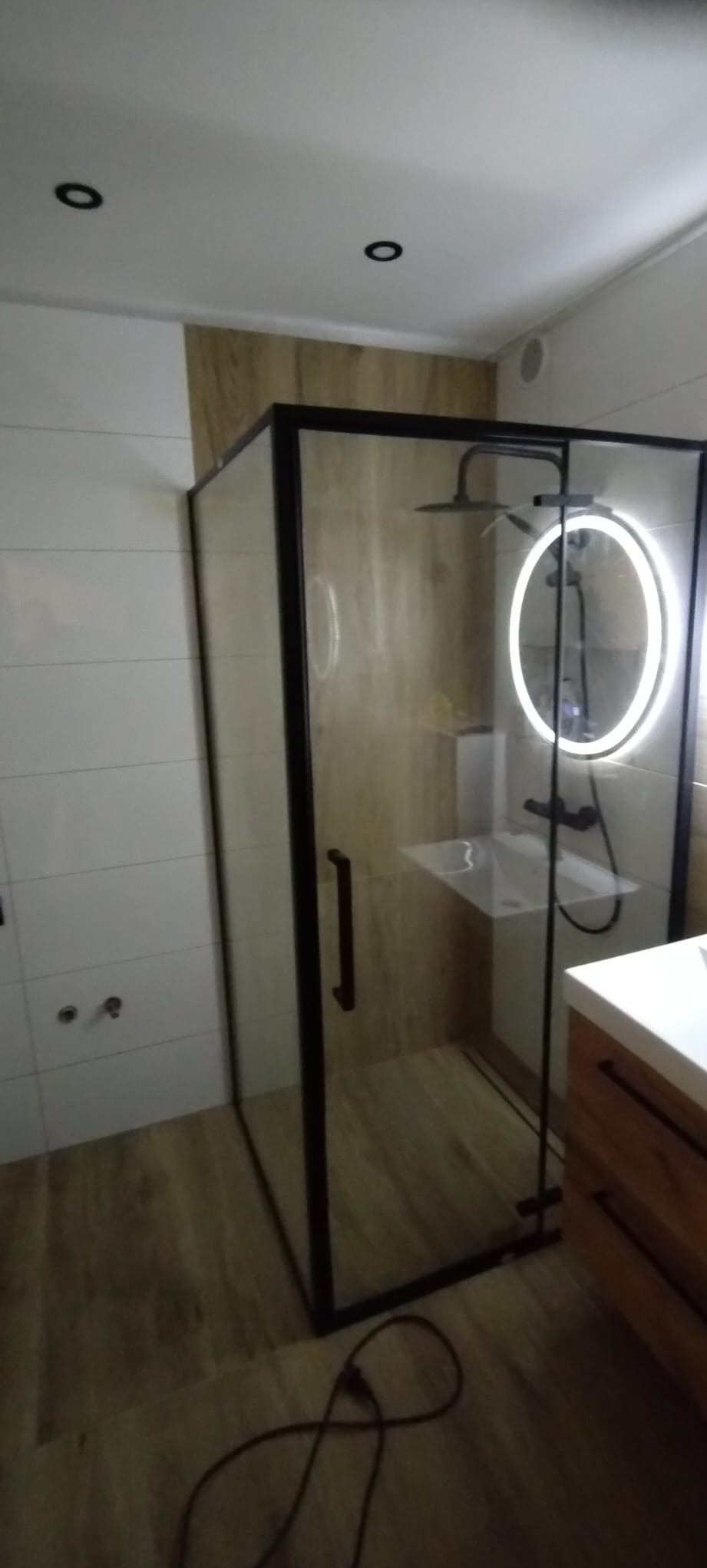 Nowoczesna kabina prysznicowa z czarnymi ramami i okrągłym lustrem z podświetleniem LED, drewnopodobne płytki na ścianie i podłodze, minimalistyczny design.