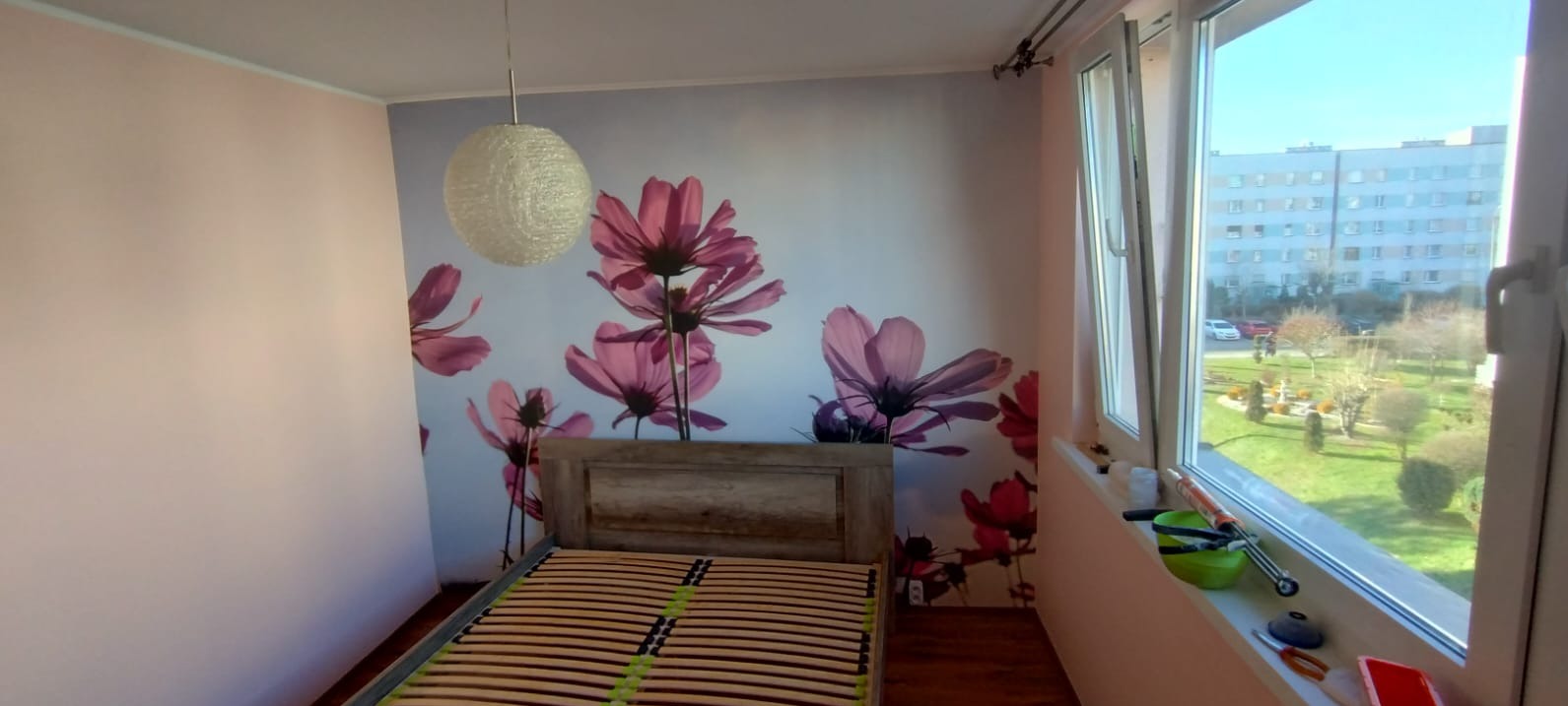 Sypialnia po remoncie: ściana z fototapetą w kwiaty, drewniane łóżko z listwami, okno z widokiem na zieleń. Na parapecie akcesoria malarskie.
