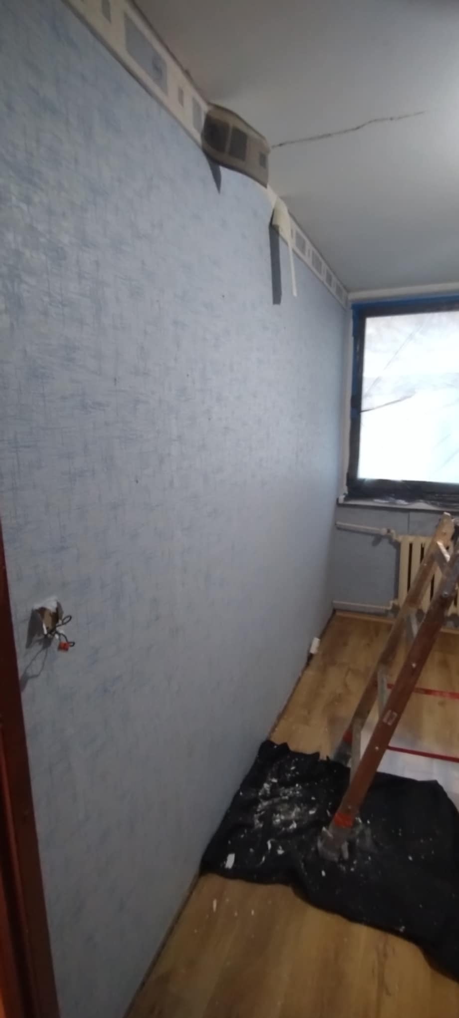 Niebieska ściana z fakturą, przygotowana do prac wykończeniowych. Widoczna drabina, zabezpieczona folią okno i wystające kable ze ściany.